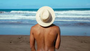 Person mit Sonnenhut, Rücken zur Kamera, steht am Strand und blickt aufs Meer.