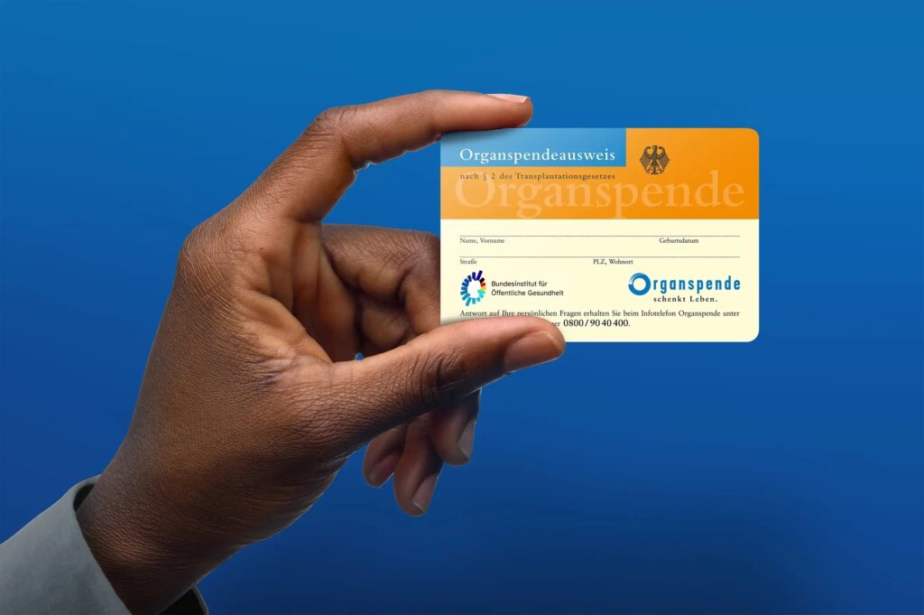 Hand hält einen orange-blauen deutschen Organspendeausweis vor blauem Hintergrund.