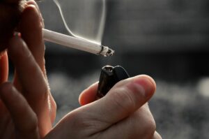Nahaufnahme: Hand hält eine brennende Zigarette und zündet sie mit einem Feuerzeug; Rauch steigt auf.
