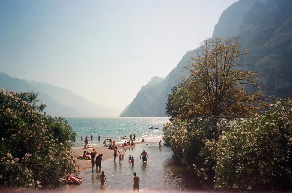 Menschen am flachen Ufer eines Bergsees, einige im Wasser, andere am Strand; Ufervegetation und steile Berge im Hintergrund.