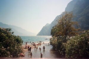Menschen am flachen Ufer eines Bergsees, einige im Wasser, andere am Strand; Ufervegetation und steile Berge im Hintergrund.