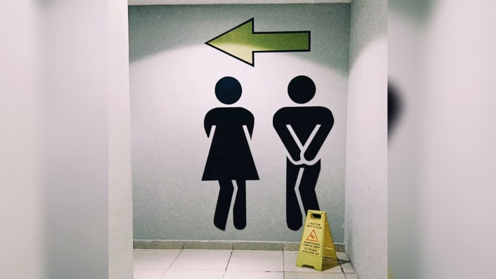 Wand mit großen Toiletten-Piktogrammen (Dame und Herr), linksweisender Pfeil oben und gelbes Warnschild für nassen Boden auf dem Fliesenboden.