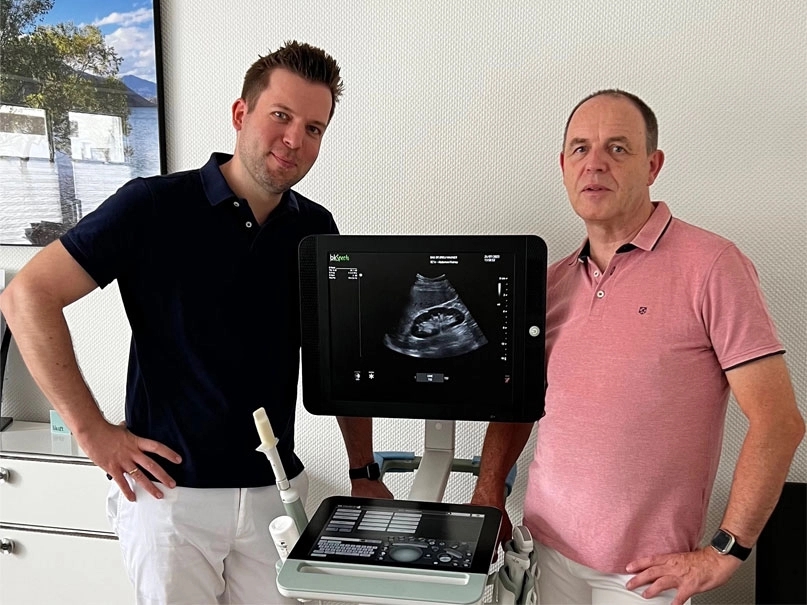 Zwei Männer stehen nebeneinander vor einem Ultraschallgerät, auf dessen Monitor ein Sonogramm angezeigt wird.