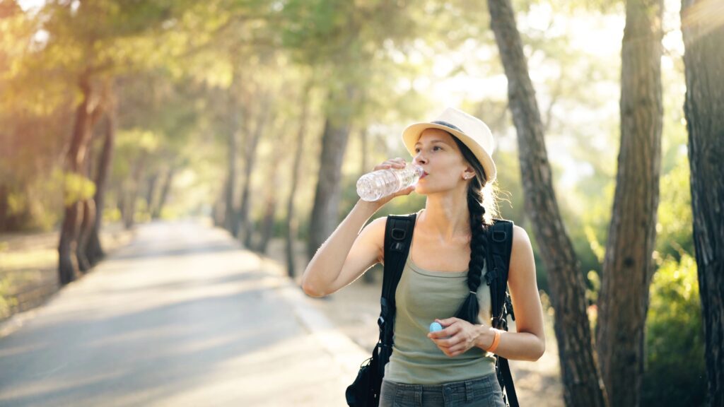 Junge Frau mit Hut und Zopf, Rucksack und Tanktop trinkt aus einer Wasserflasche auf einem sonnigen Waldweg.