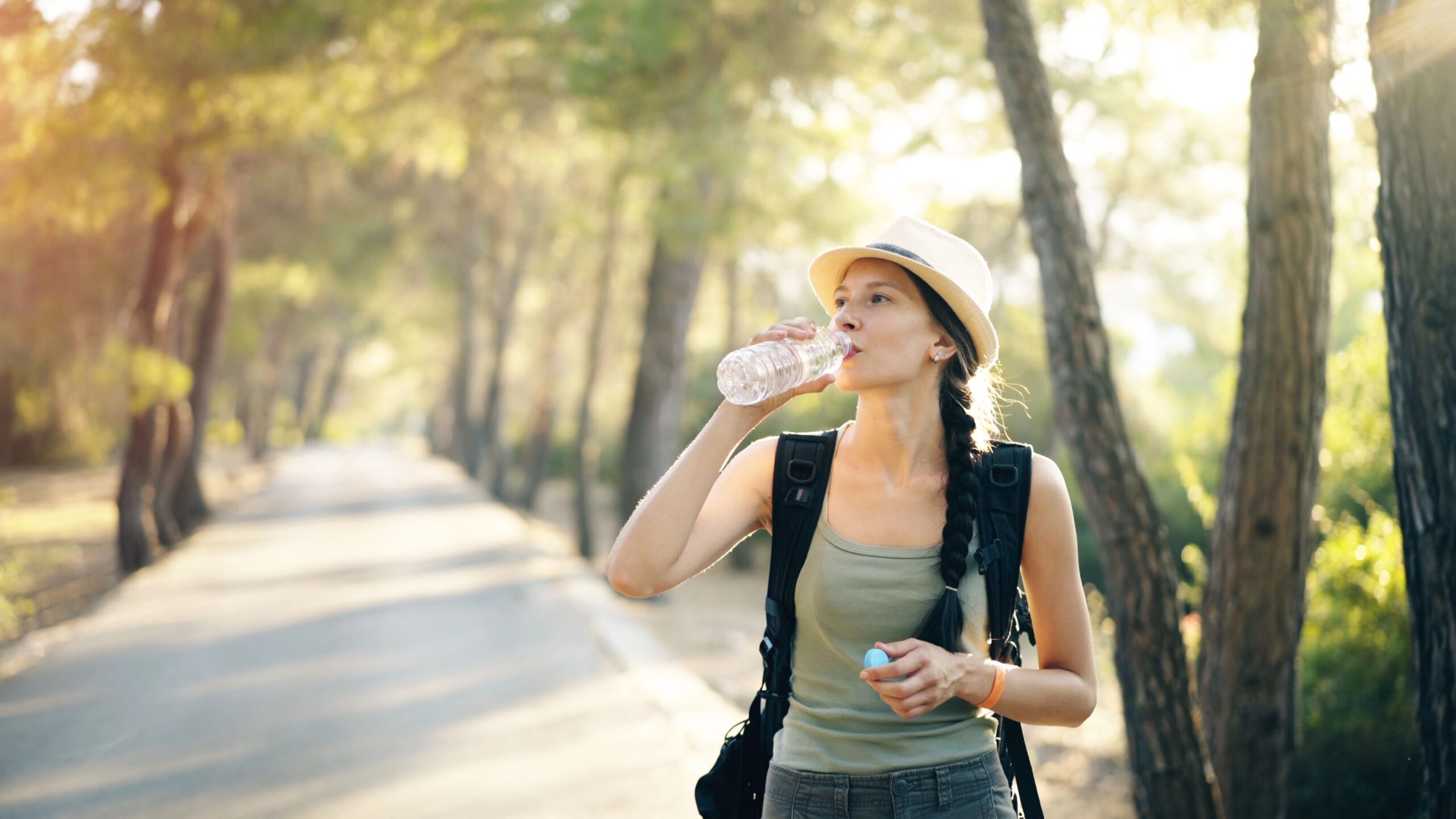 Junge Frau mit Hut und Zopf, Rucksack und Tanktop trinkt aus einer Wasserflasche auf einem sonnigen Waldweg.