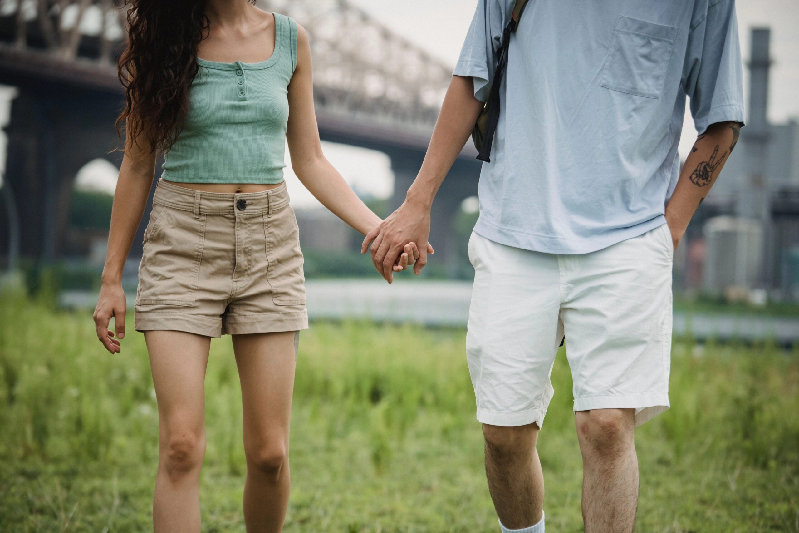 Zwei Menschen gehen nebeneinander im Freien und halten sich an den Händen, mit einer Brücke im Hintergrund.