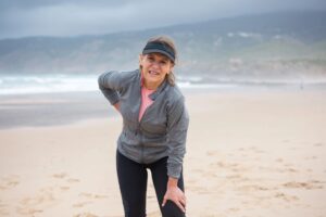 Frau in Sportkleidung mit Visor steht am Strand und hält sich die untere Rückenregion, im Hintergrund Meer und Dünen.
