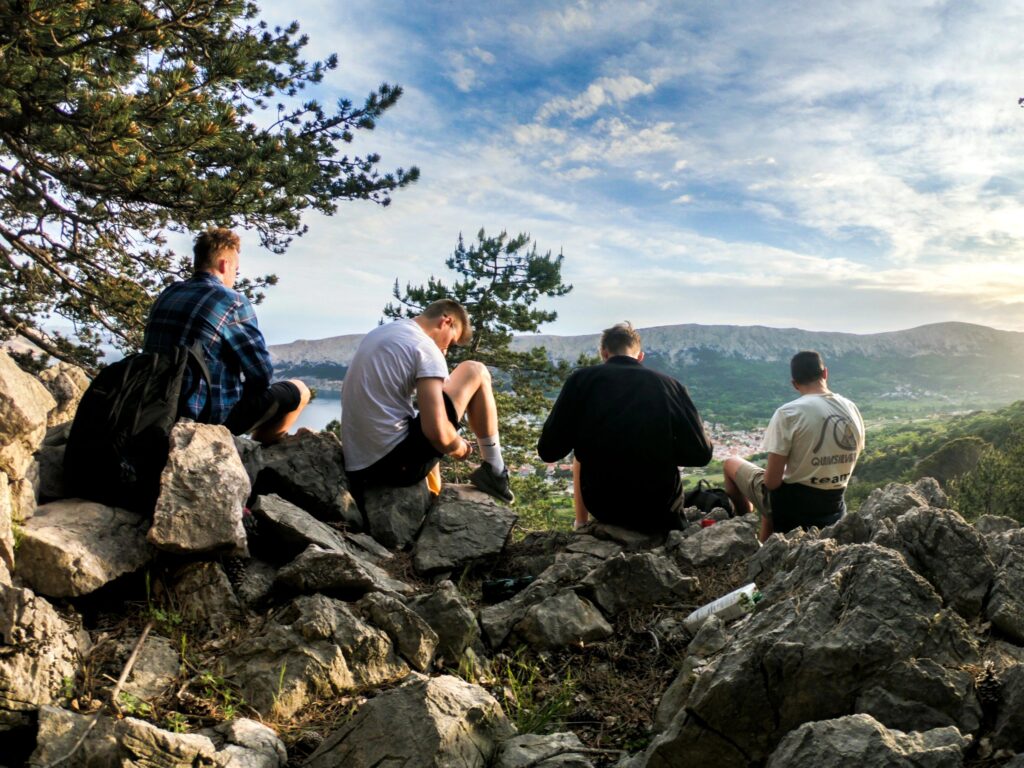 Vier Personen sitzen auf Felsen und schauen auf ein Tal mit Hügeln und einem Gewässer unter blauem Himmel.