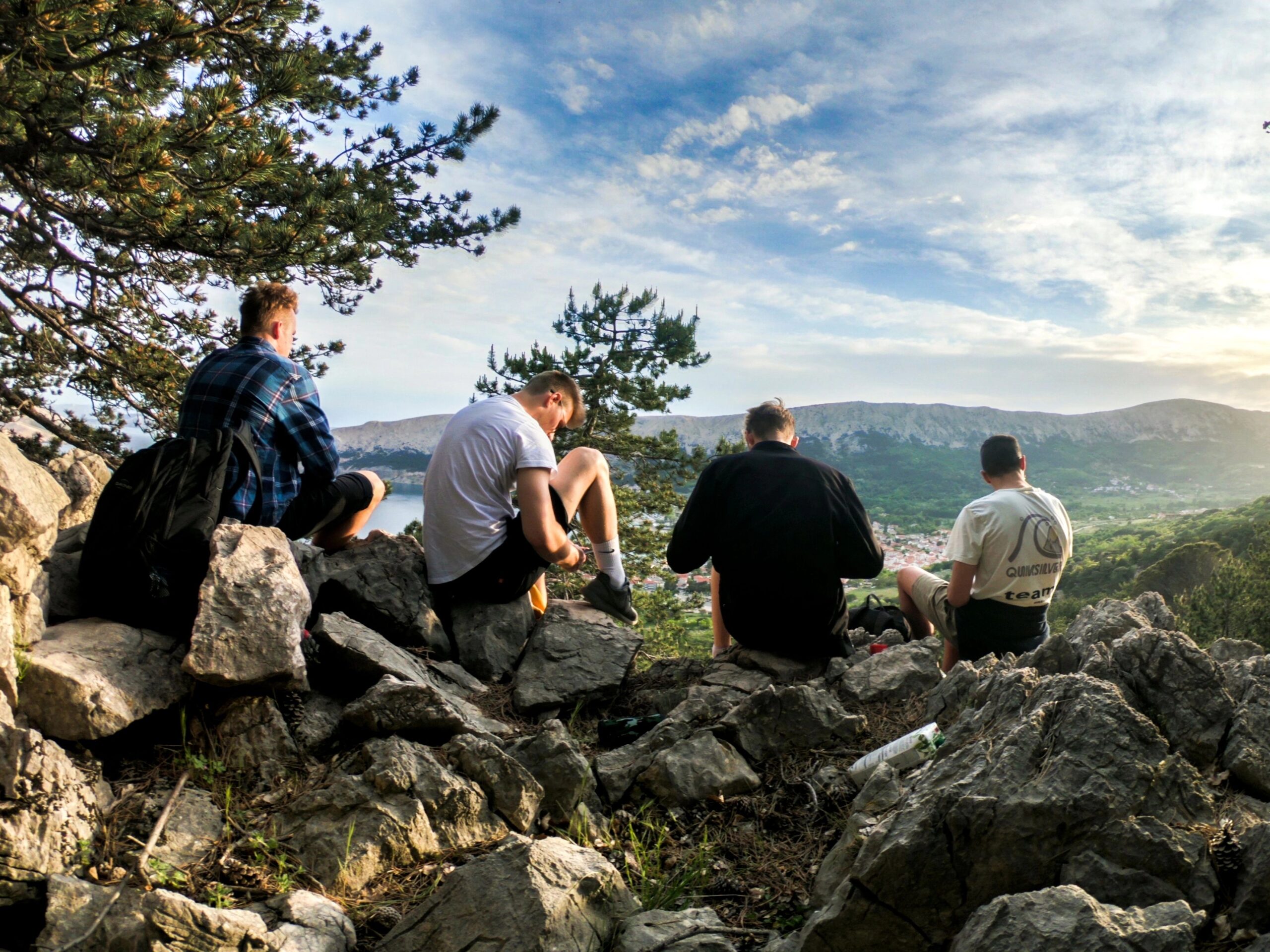 Vier Personen sitzen auf Felsen und schauen auf ein Tal mit Hügeln und einem Gewässer unter blauem Himmel.