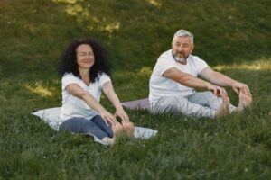 Älteres Mann und Frau sitzen auf Yogamatten auf einer Wiese und dehnen sich nach vorne, wobei sie ihre Füße halten.