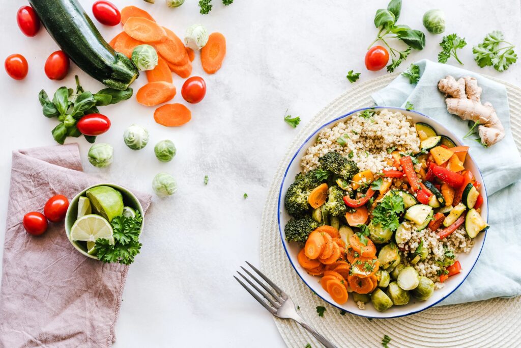 Schüssel mit Quinoa, Brokkoli, Karotten, Rosenkohl und anderem Gemüse, dazu Tomaten, Limette und frische Kräuter auf hellem Untergrund.